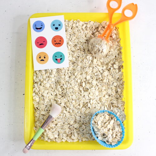 This image shows a sensory play activity setup with a yellow tray filled with dry oats. A pair of orange scoop scissors and a small brush are included, along with a blue cupcake liner filled with oats. On the left side, there is a card featuring various colorful emoji faces expressing different emotions, such as happy, sad, angry, and surprised. This setup is likely intended for a tactile sensory experience, helping children explore textures and emotions through play.