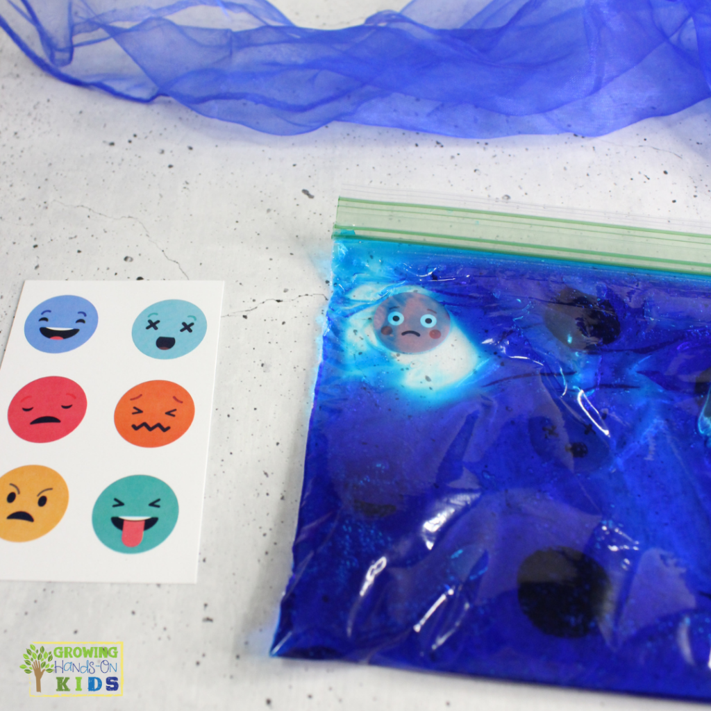 A DIY emotions sensory bag filled with blue gel, featuring a sad face emoji inside, next to a card with various emotion face stickers, including happy, sad, angry, and surprised expressions. A blue scarf is visible in the background.