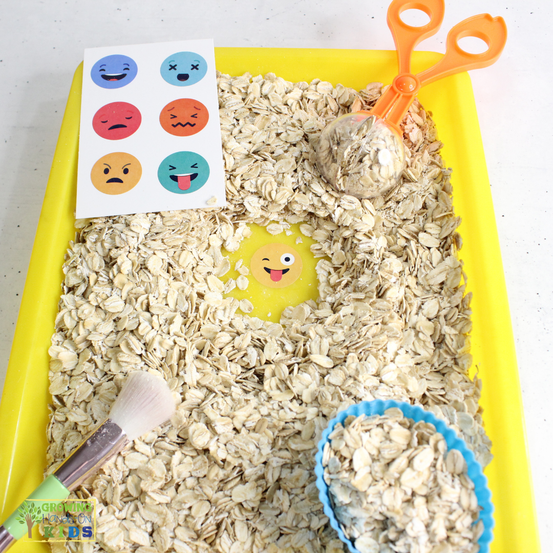 This image shows a sensory play activity setup with a yellow tray filled with dry oats. A pair of orange scoop scissors and a small brush are included, along with a blue cupcake liner filled with oats. On the left side, there is a card featuring various colorful emoji faces expressing different emotions, such as happy, sad, angry, and surprised. This setup is intended for a tactile sensory experience, helping children explore textures and emotions through play.