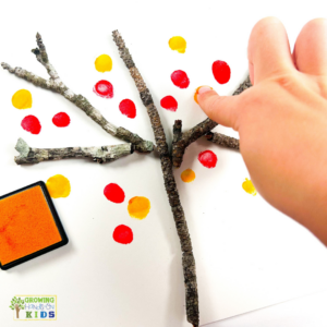 A child's hand pressing a fingertip onto white paper, creating red and yellow fingerprint leaves around twigs arranged as tree branches for a fall fingerprint tree craft.