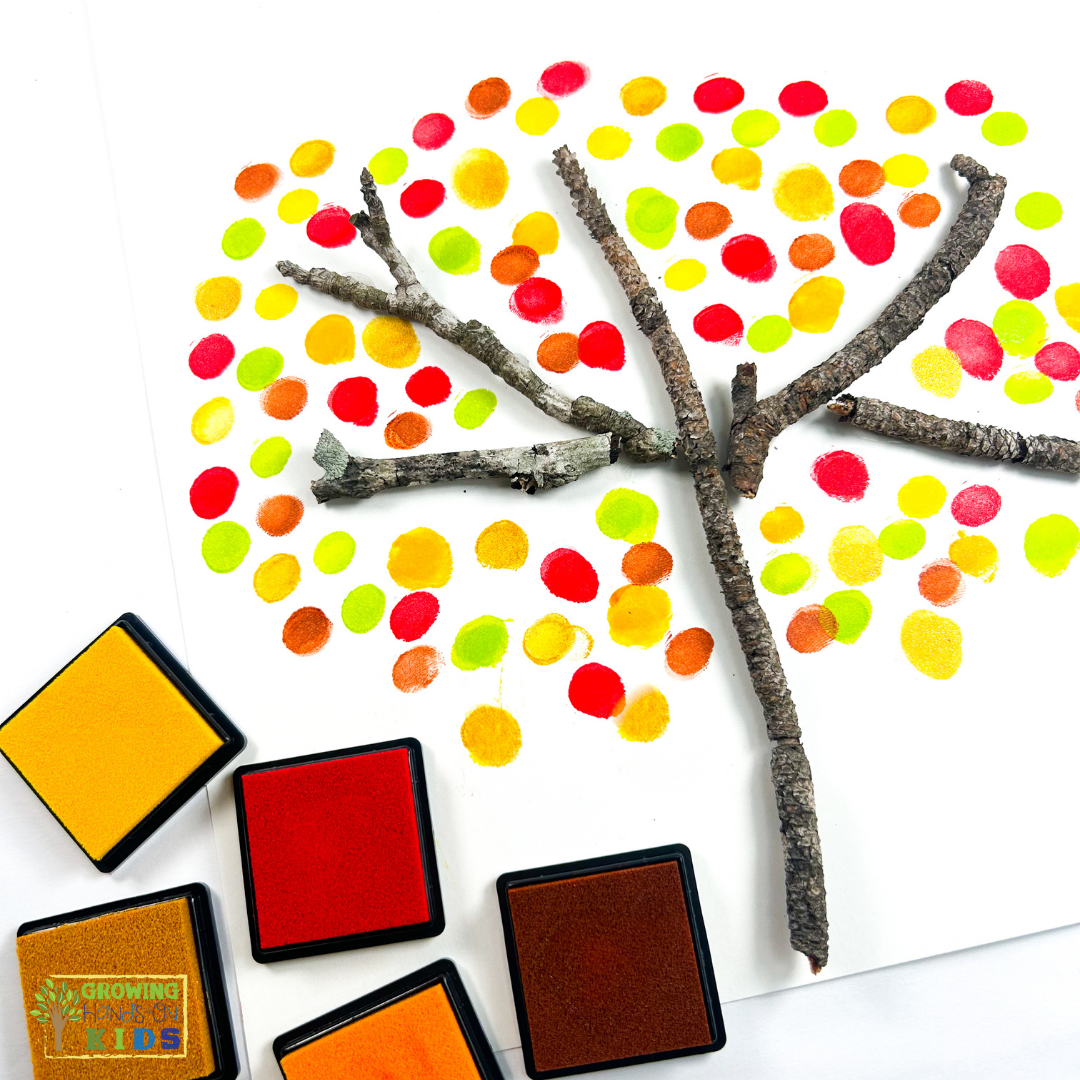 A fall craft featuring real twigs arranged as branches on white paper, surrounded by colorful fingerprint dots in red, orange, yellow, and green. Ink pads in fall colors are placed next to the artwork.