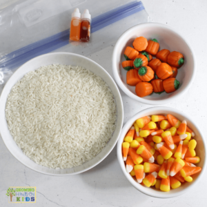 Ingredients for a candy corn sensory bin displayed on a table, including a bowl of white rice, a bowl of candy corn, a bowl of candy pumpkins, two small bottles of food dye, and a large Ziploc bag.