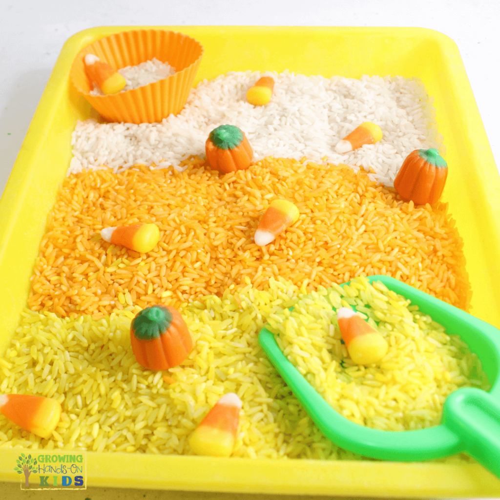 Yellow sensory bin filled with layers of white, orange, and yellow rice, decorated with candy corn and candy pumpkins, alongside a green scoop and an orange silicone cup for scooping.