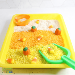 Yellow sensory bin filled with layers of white, orange, and yellow rice, decorated with candy corn and candy pumpkins, alongside a green scoop and an orange silicone cup for scooping.