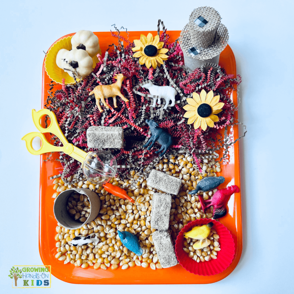 Farm animal sensory bin setup featuring plastic farm animals, corn kernels, sunflowers, shredded paper, and small toys on an orange tray for hands-on sensory play