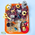 Farm animal sensory bin setup featuring plastic farm animals, corn kernels, sunflowers, shredded paper, and small toys on an orange tray for hands-on sensory play.