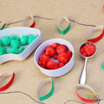 Christmas-themed activity setup featuring red and green glitter pom poms in white bowls, a metal scoop with red pom poms, and painted toilet paper roll strips resembling Christmas lights glued onto cardboard with a drawn string connecting them.