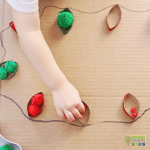 Child picking up a pom-pom in a Christmas-themed activity setup featuring red and green glitter pom poms in white bowls, a metal scoop with red pom poms, and painted toilet paper roll strips resembling Christmas lights glued onto cardboard with a drawn string connecting them.